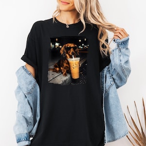 May include: Black t-shirt featuring a dachshund wearing sunglasses, holding a coffee drink with a straw. The dog is sitting on a sidewalk. The image is a graphic design.