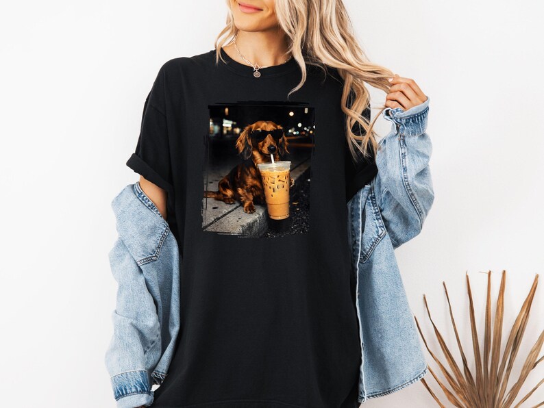 May include: Black t-shirt featuring a dachshund wearing sunglasses, holding a coffee drink with a straw. The dog is sitting on a sidewalk. The image is a graphic design.