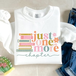 May include: White sweatshirt with the text "just one more chapter" in colourful letters, with a stack of books in the background.
