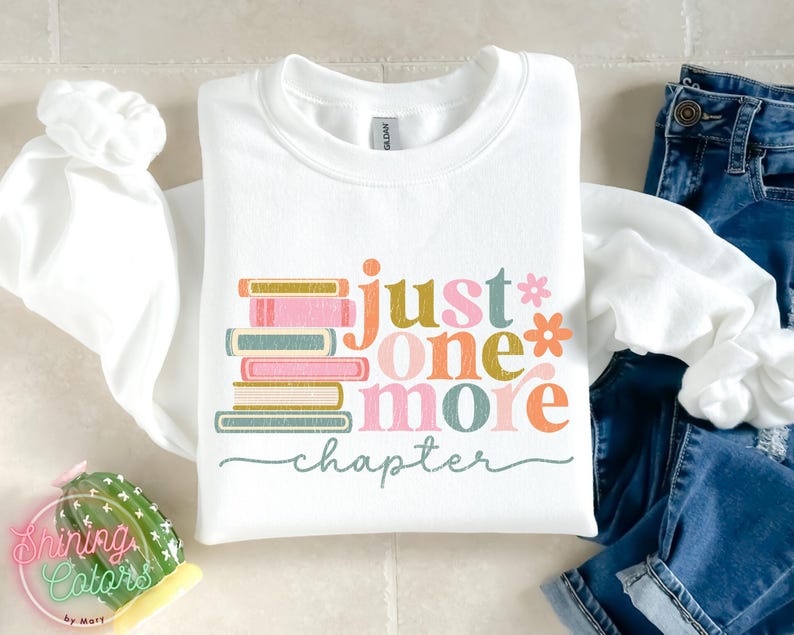 May include: White sweatshirt with the text "just one more chapter" in colourful letters, with a stack of books in the background.