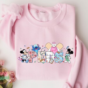 May include: Pink sweatshirt with a graphic of Stitch, Mickey Mouse, and Baymax dressed as nurses. The graphic includes the word "PEDS" in a colorful, medical-themed design.