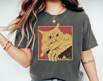 May include: Dark grey t-shirt featuring a cartoon cat illustration. The cat, in shades of yellow and brown, is set against a red and yellow background. The cat is holding a finger in its mouth. The shirt is a casual style.