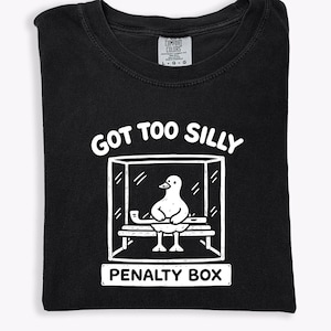 May include: Black t-shirt featuring a white graphic of a duck in a penalty box. The text "GOT TOO SILLY" is above the box, and "PENALTY BOX" is below. The shirt is folded and made of cotton.