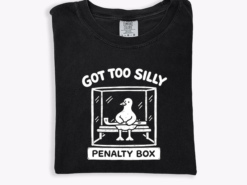 May include: Black t-shirt featuring a white graphic of a duck in a penalty box. The text "GOT TOO SILLY" is above the box, and "PENALTY BOX" is below. The shirt is folded and made of cotton.