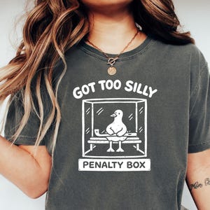 May include: Dark grey t-shirt featuring a white graphic of a duck in a penalty box. The text "GOT TOO SILLY" is above the box, and "PENALTY BOX" is below. The shirt has a relaxed fit.