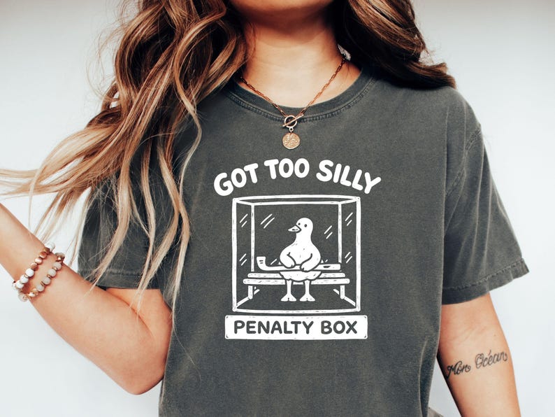 May include: Dark grey t-shirt featuring a white graphic of a duck in a penalty box. The text "GOT TOO SILLY" is above the box, and "PENALTY BOX" is below. The shirt has a relaxed fit.