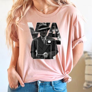 Puede incluir: Camiseta rosa claro con una imagen en blanco y negro de un hombre con traje y sombrero, con la palabra "VAULT" superpuesta. La imagen est&aacute; centrada en la camiseta.