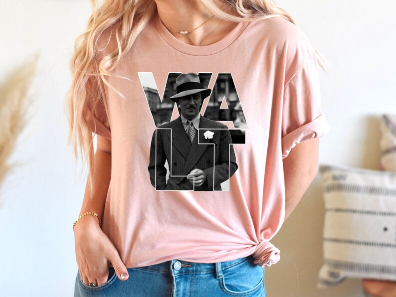 Puede incluir: Camiseta rosa claro con una imagen en blanco y negro de un hombre con traje y sombrero, con la palabra "VAULT" superpuesta. La imagen est&aacute; centrada en la camiseta.