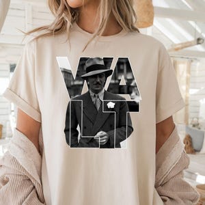 Puede incluir: Camiseta beige con una imagen en blanco y negro de un hombre con traje y sombrero, con la palabra "VAULT" en letras blancas. La camiseta tiene mangas cortas y un corte holgado.
