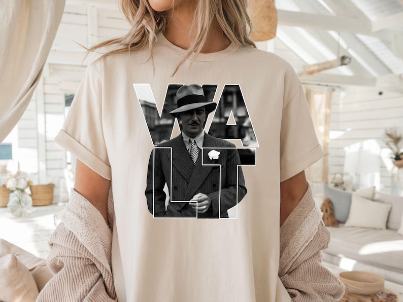 Puede incluir: Camiseta beige con una imagen en blanco y negro de un hombre con traje y sombrero, con la palabra "VAULT" en letras blancas. La camiseta tiene mangas cortas y un corte holgado.