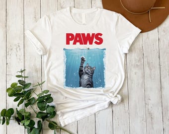 May include: White t-shirt featuring a parody of the movie "Jaws" with the word "PAWS" in red block letters above an illustration of a cat reaching out of the water. The cat is gray and white with its paw extended.