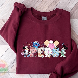 May include: Burgundy sweatshirt with a graphic of Disney characters dressed as nurses. The characters include Stitch, Mickey Mouse, Minnie Mouse, and Baymax. The text "Peds" is written in a colorful font.