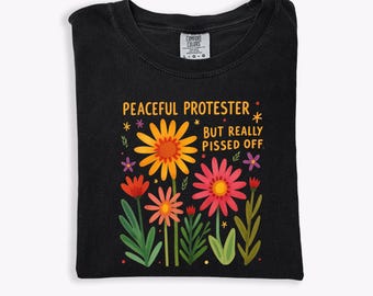 Puede incluir: Camiseta negra con un diseño floral y el texto "PEACEFUL PROTESTER BUT REALLY PISSED OFF". El gráfico presenta flores coloridas en tonos de amarillo, naranja, rosa y rojo, con hojas y tallos verdes. La camiseta está doblada.