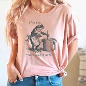 May include: Pink t-shirt with a humorous graphic of a frog wearing an apron and stirring a pot. The text above the frog reads "That's it." and below it says "You're going in the SOUP."