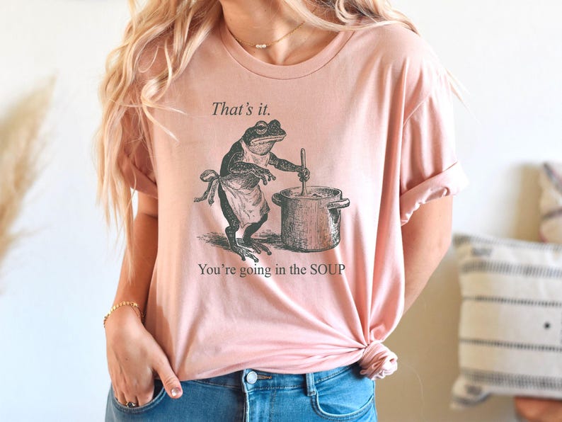 May include: Pink t-shirt with a humorous graphic of a frog wearing an apron and stirring a pot. The text above the frog reads "That's it." and below it says "You're going in the SOUP."