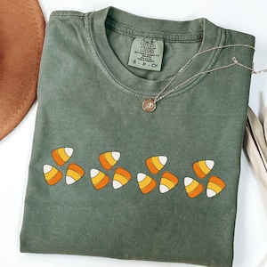 May include: A sage green t-shirt with a candy corn graphic. The shirt has a crew neck and short sleeves. A gold necklace with a pendant is draped across the shirt. A brown hat and dried plant are in the background.