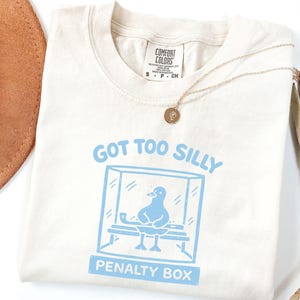 May include: Cream-coloured t-shirt featuring a light blue graphic of a bird in a penalty box, with the text "GOT TOO SILLY PENALTY BOX". The shirt has a round neckline and short sleeves. A gold necklace is draped across the shirt.