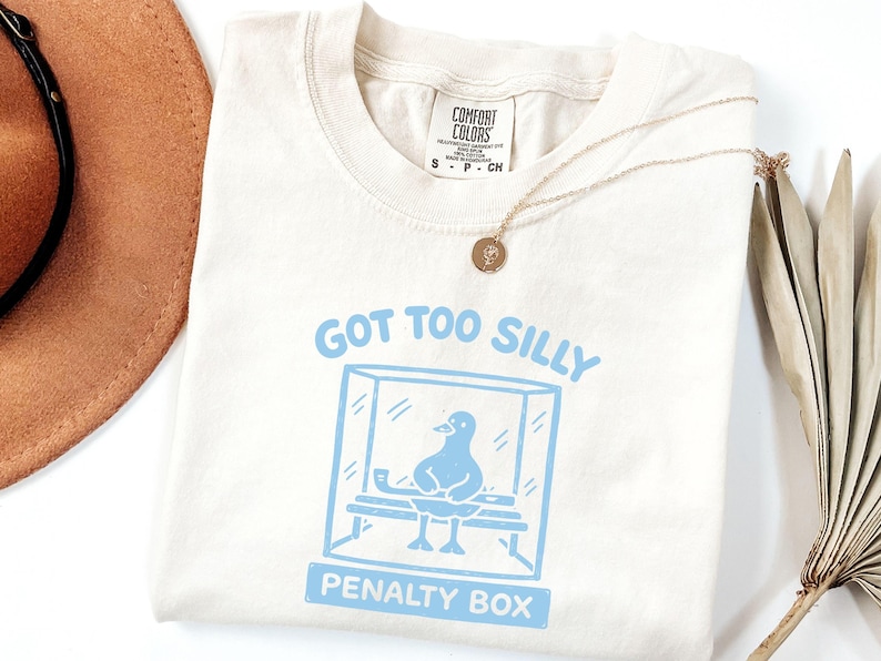 May include: Cream-coloured t-shirt featuring a light blue graphic of a bird in a penalty box, with the text "GOT TOO SILLY PENALTY BOX". The shirt has a round neckline and short sleeves. A gold necklace is draped across the shirt.