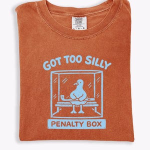 May include: Burnt orange t-shirt featuring a light blue graphic of a duck in a penalty box, with the text "GOT TOO SILLY" above and "PENALTY BOX" below. The shirt has a crew neck and a Comfort Colors label.