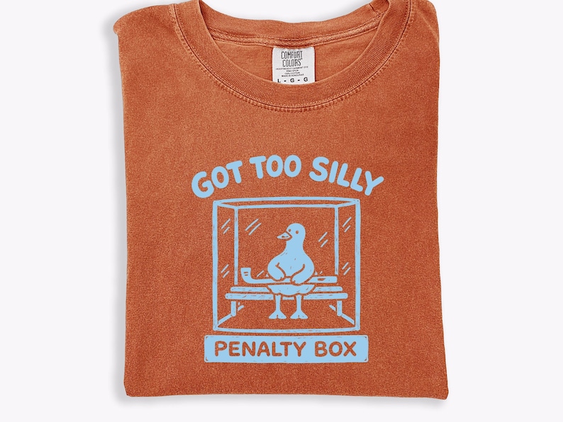 May include: Burnt orange t-shirt featuring a light blue graphic of a duck in a penalty box, with the text "GOT TOO SILLY" above and "PENALTY BOX" below. The shirt has a crew neck and a Comfort Colors label.