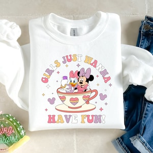 Puede incluir: Sudadera blanca con un gr&aacute;fico rosa, amarillo y azul de Minnie Mouse y Daisy Duck en una taza de t&eacute;. El texto "Girls Just Wanna Have Fun!" est&aacute; impreso en una fuente retro.