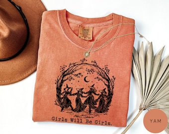 May include: A coral-colored t-shirt with a round neck and a graphic of four witches dancing in a circle under a crescent moon. The text "Girls Will Be Girls" is printed below the graphic. A gold necklace and a brown hat are also visible.