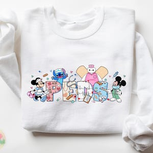 May include: White sweatshirt with a colorful graphic of cartoon characters dressed as nurses. The graphic includes Mickey Mouse, Stitch, Baymax, and the word "Peds".