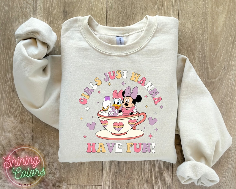 Puede incluir: Una sudadera de color beige claro con un estampado gr&aacute;fico de Mickey Mouse y Daisy Duck en una taza de t&eacute;. El texto "Girls Just Wanna Have Fun!" est&aacute; impreso en una fuente de arco&iacute;ris alrededor de la taza de t&eacute;.