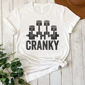 May include: White t-shirt featuring a black graphic of four engine pistons and the word "CRANKY" in a distressed font. The shirt is made of a soft material and has a classic crew neck design. A casual and comfortable apparel item.