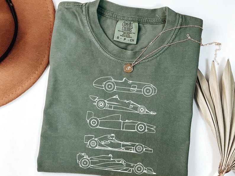 K&ouml;nnte beinhalten: Olivgr&uuml;nes T-Shirt mit einer wei&szlig;en Strichzeichnung von f&uuml;nf Rennwagen. Das T-Shirt hat einen Rundhalsausschnitt und kurze &Auml;rmel. Eine goldene Halskette mit einem runden Anh&auml;nger liegt &uuml;ber dem T-Shirt.