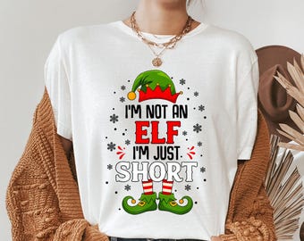 May include: White t-shirt with the text "I'M NOT AN ELF I'M JUST SHORT" in black, red, and white. The design includes an elf hat, snowflakes, and elf shoes. The shirt is a festive holiday item.
