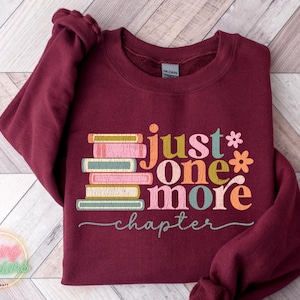 May include: Burgundy sweatshirt featuring a stack of colourful books and the text "just one more chapter" in a retro font.