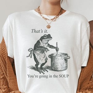 May include: White t-shirt featuring a vintage-style illustration of a frog wearing an apron and stirring a pot. The text "That's it. You're going in the SOUP" is printed above and below the frog graphic.