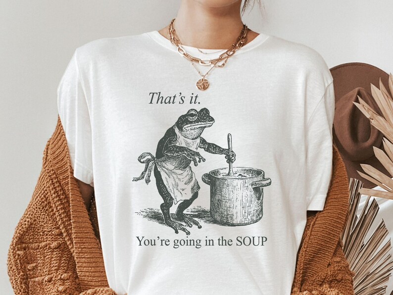 May include: White t-shirt featuring a vintage-style illustration of a frog wearing an apron and stirring a pot. The text "That's it. You're going in the SOUP" is printed above and below the frog graphic.