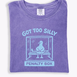 May include: A purple t-shirt featuring a light blue graphic with the text "GOT TOO SILLY" and a cartoon duck in a penalty box. The shirt has a crew neck and a Comfort Colors label.