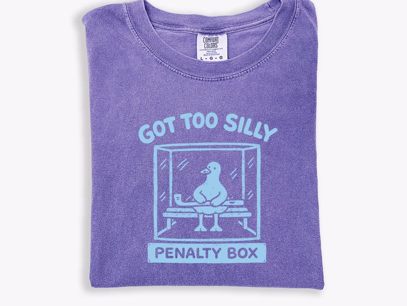 May include: A purple t-shirt featuring a light blue graphic with the text "GOT TOO SILLY" and a cartoon duck in a penalty box. The shirt has a crew neck and a Comfort Colors label.