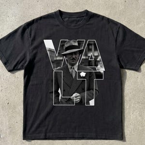Puede incluir: Camiseta negra con una imagen en blanco y negro de un hombre con traje y sombrero, superpuesta con la palabra "WALT" en letras blancas. El hombre lleva un traje y corbata.
