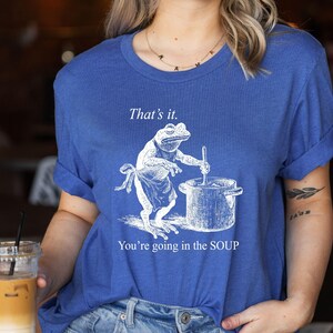 May include: Blue t-shirt featuring a frog chef stirring a pot, with the text "That's it. You're going in the SOUP." The frog wears an apron. The shirt is a casual, unisex style.