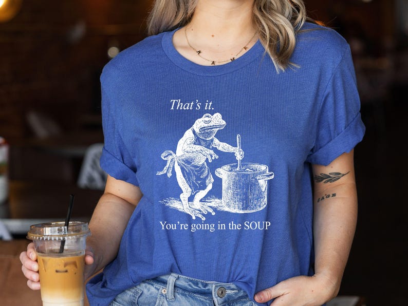 May include: Blue t-shirt featuring a frog chef stirring a pot, with the text "That's it. You're going in the SOUP." The frog wears an apron. The shirt is a casual, unisex style.