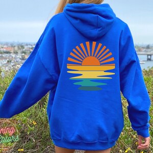 May include: A royal blue hoodie with a retro sunset graphic on the back. The graphic features a yellow sun with orange rays and a blue, green, and yellow reflection in the water.