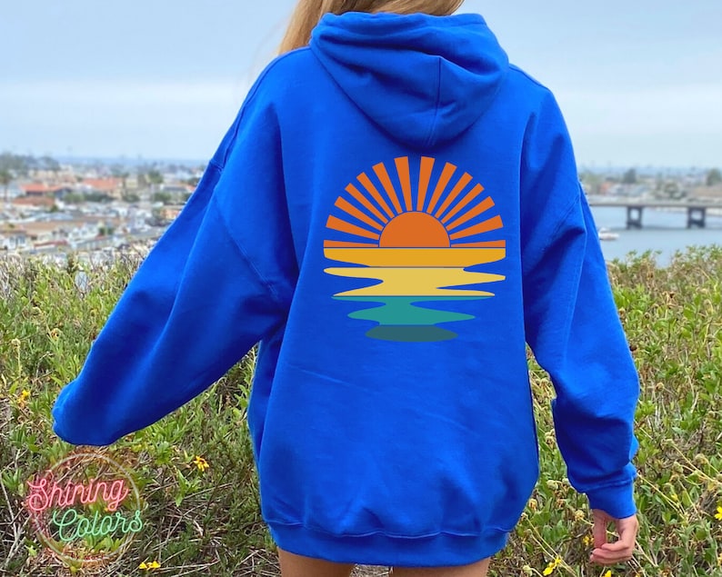 May include: A royal blue hoodie with a retro sunset graphic on the back. The graphic features a yellow sun with orange rays and a blue, green, and yellow reflection in the water.