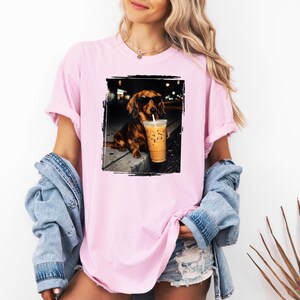 May include: Pink t-shirt featuring a framed image of a dachshund wearing sunglasses and holding an iced coffee. The dog is brown with black accents. The shirt has short sleeves and a relaxed fit.