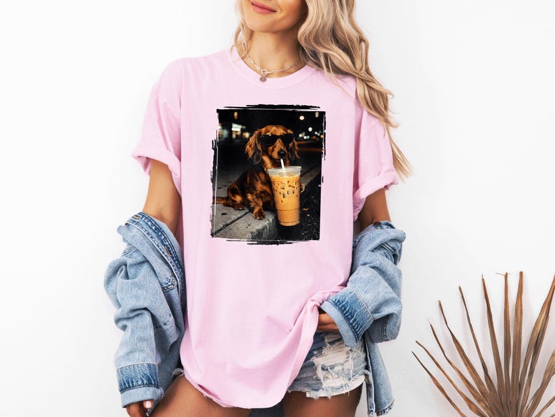 May include: Pink t-shirt featuring a framed image of a dachshund wearing sunglasses and holding an iced coffee. The dog is brown with black accents. The shirt has short sleeves and a relaxed fit.