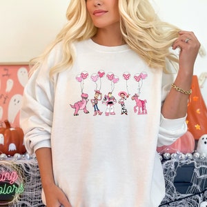 May include: White sweatshirt with a pink and white graphic of Toy Story characters holding pink balloons. The characters include Rex, Woody, Buzz Lightyear, Jessie, and Bullseye.