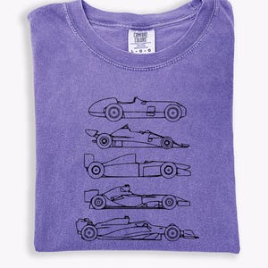 K&ouml;nnte beinhalten: Ein lila Comfort Colors T-Shirt mit einer schwarzen Strichzeichnung von f&uuml;nf Rennwagen. Das T-Shirt hat einen Rundhalsausschnitt und ist gefaltet. Das Etikett lautet "Comfort Colors" und enth&auml;lt Gr&ouml;&szlig;enangaben.