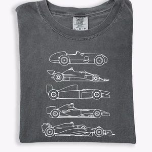 K&ouml;nnte beinhalten: Dunkelgraues Comfort Colors T-Shirt mit einer wei&szlig;en Strichzeichnung von f&uuml;nf verschiedenen Rennwagen. Die Autos sind im Profil dargestellt und zeigen ihre unterschiedlichen Designs. Das Shirt hat einen Rundhalsausschnitt und ein Etikett mit der Aufschrift "Comfort Colors".