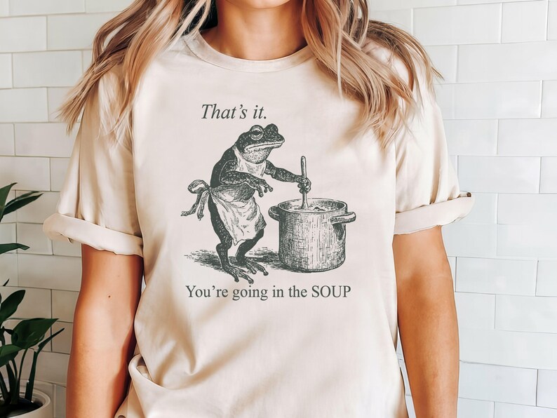 May include: Beige t-shirt featuring a black and white illustration of a frog wearing an apron and stirring a pot. Text above reads "That's it." and below "You're going in the SOUP."