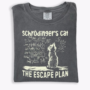 May include: Dark gray t-shirt featuring the text "Schrödinger's Cat" above a cat illustration and the words "The Escape Plan." The design includes scientific equations and diagrams in a white font.
