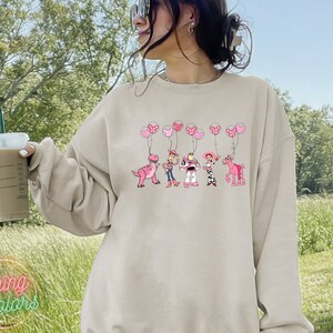 May include: A light beige sweatshirt with a graphic print of Toy Story characters holding pink balloons. The characters include Woody, Buzz Lightyear, Jessie, Rex, and a pink horse.
