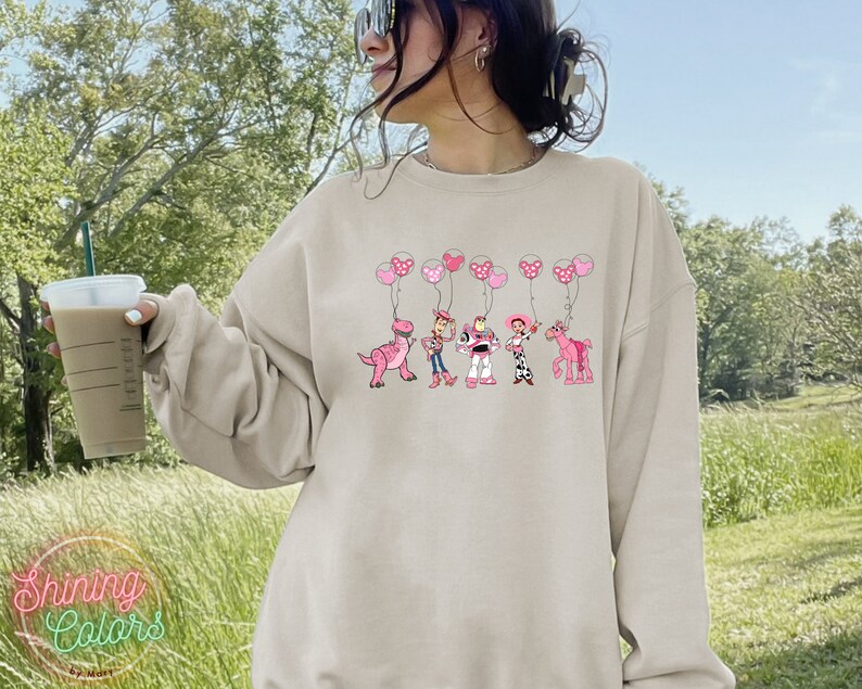 May include: A light beige sweatshirt with a graphic print of Toy Story characters holding pink balloons. The characters include Woody, Buzz Lightyear, Jessie, Rex, and a pink horse.